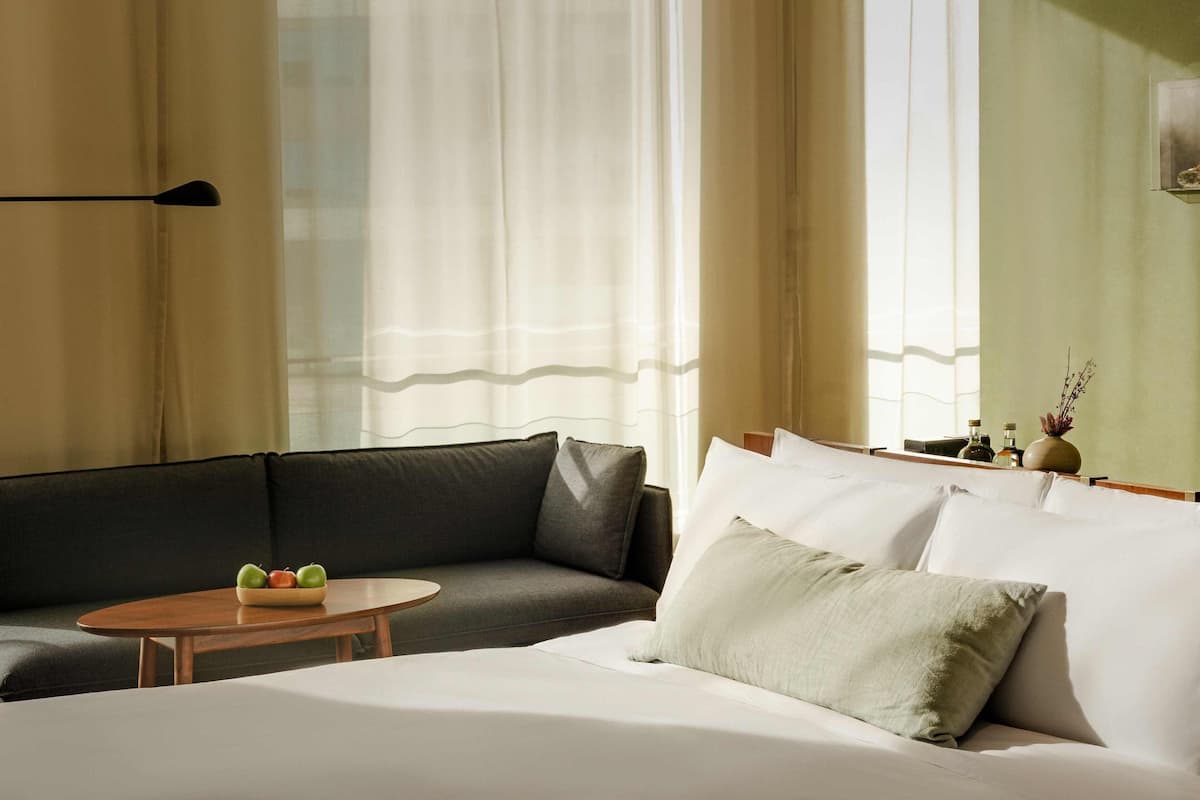A modern hotel room with a bed, a sofa, a small table with fruit, and a vase with branches.