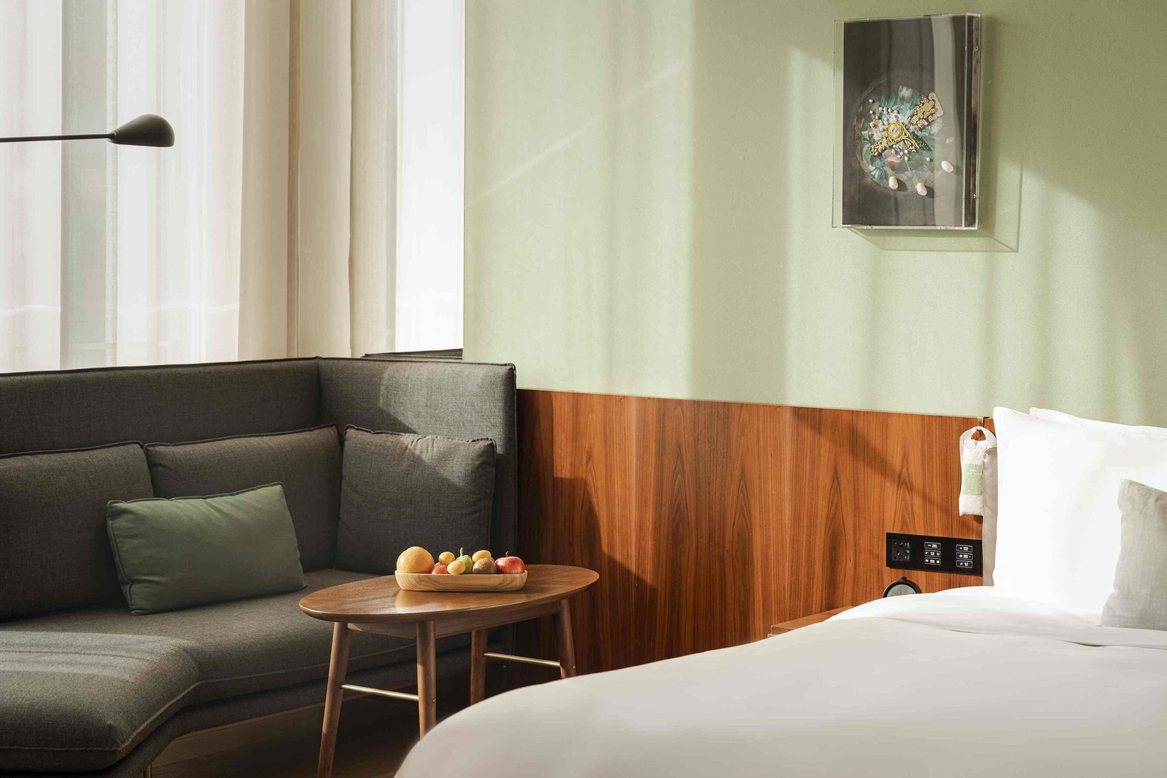 Ein Hotelzimmer mit einer Couch, einem kleinen runden Tisch mit Obst, einem Bett und einem Gemälde an der Wand.