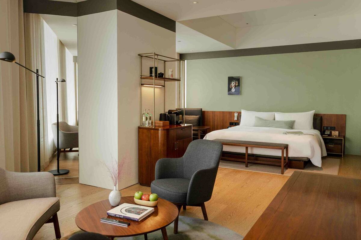 A modern hotel room with a bed, a desk, a chair, a coffee table, and a bookshelf.