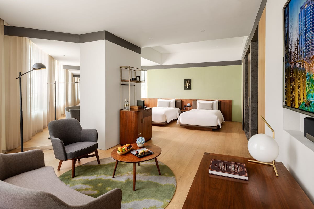A modern hotel room with a sofa, a round table with a fruit bowl, a bed with white linens, a wooden cabinet, and a painting on the wall.