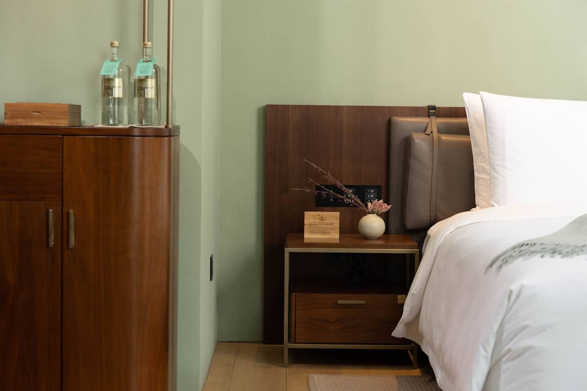 A neatly arranged hotel room with a wooden wardrobe, a bedside table with a vase of dried branches, and a bed with a white pillow and a brown headboard.