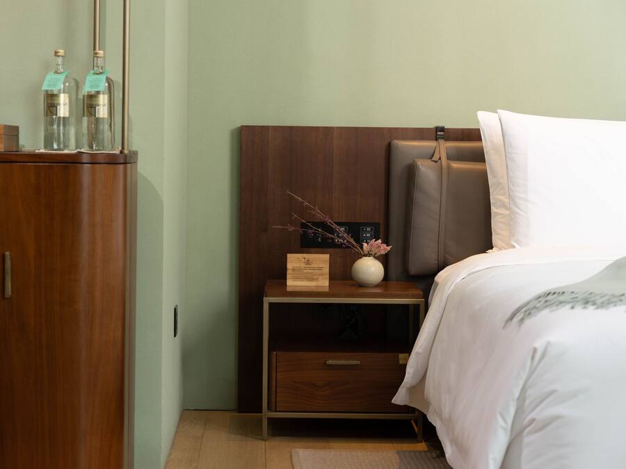 A neatly arranged hotel room with a wooden wardrobe, a bedside table with a vase of dried branches, and a bed with a white pillow and a brown headboard.