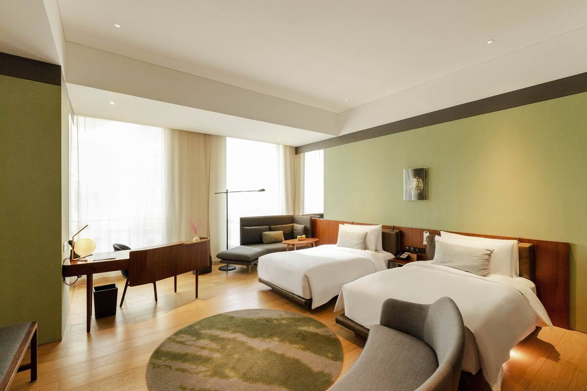 A hotel room with two beds, a desk, a sofa, and a large window.