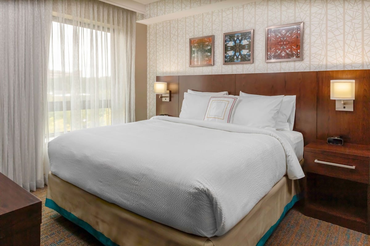 Una habitación de hotel con una cama grande, una mesita de noche, una lámpara y una ventana con cortinas.