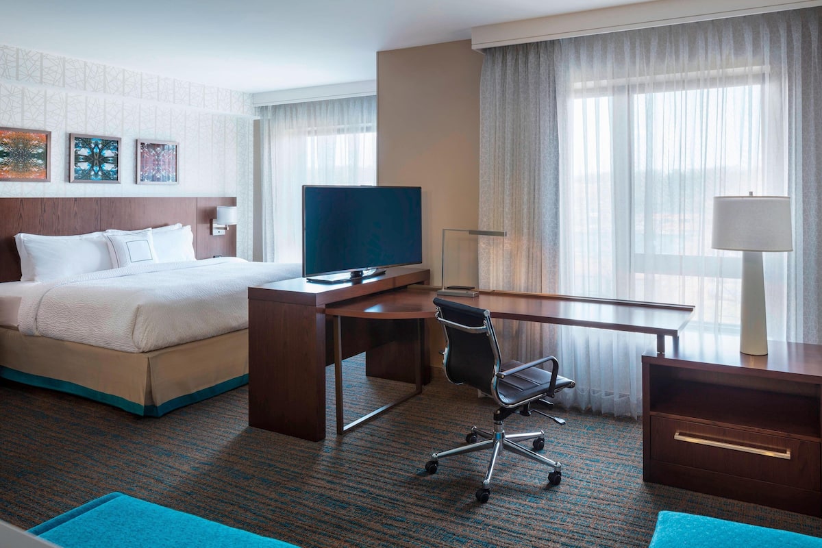Habitación de hotel con cama, escritorio, silla, televisor, lámpara y una obra de arte en la pared.