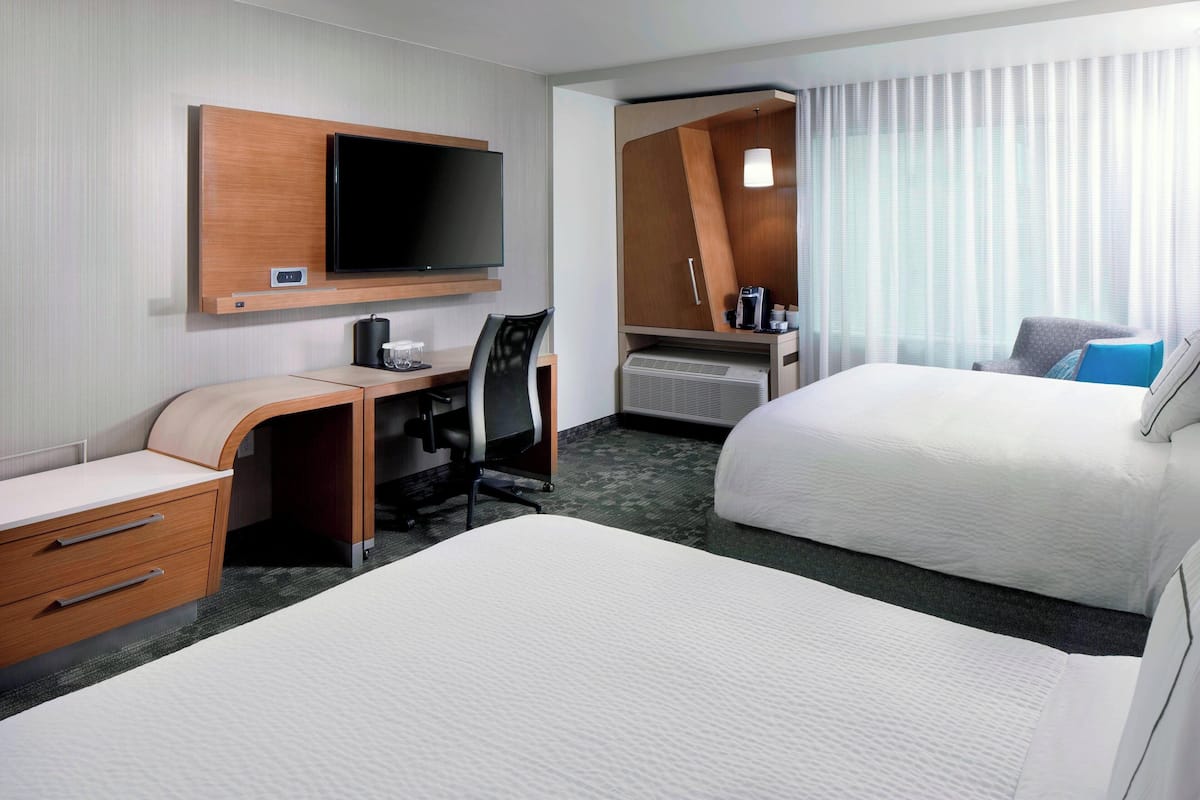 A hotel room with two beds, a desk with a chair, a television, and a large window with curtains.