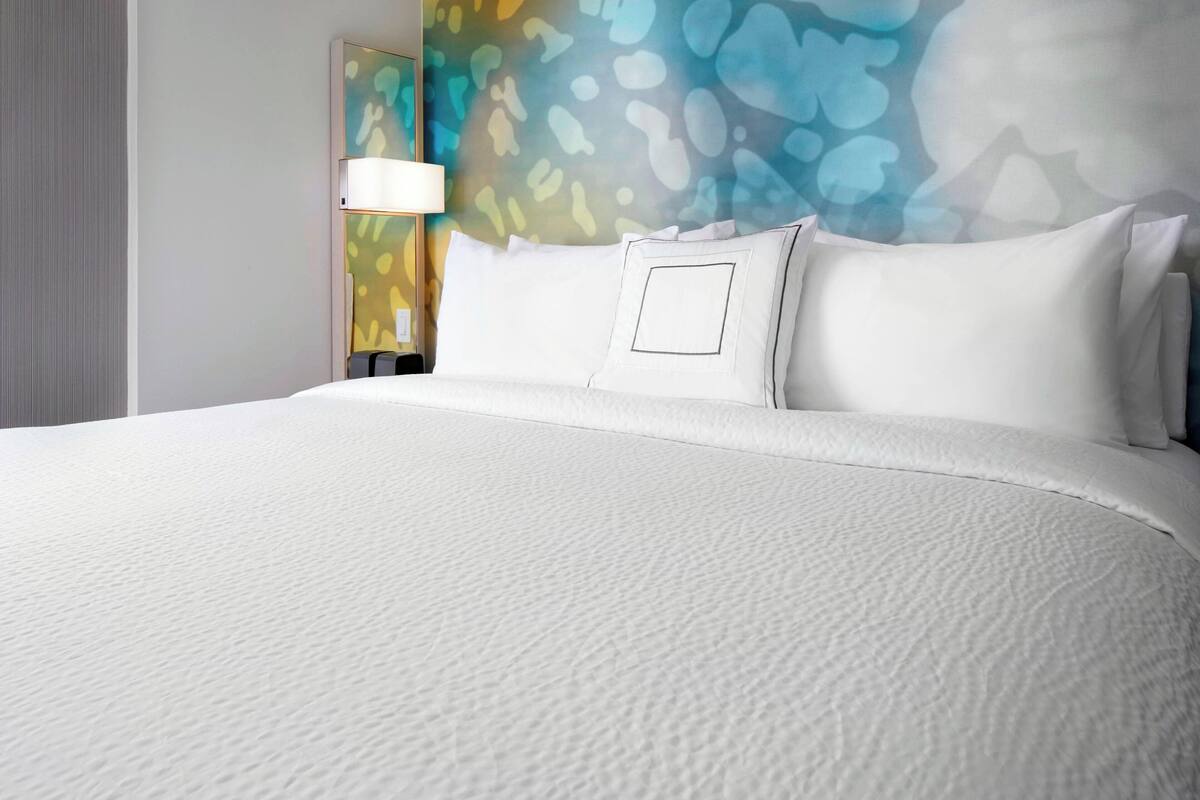 A hotel room with a large bed, a nightstand with a lamp, and a colorful abstract wall mural.