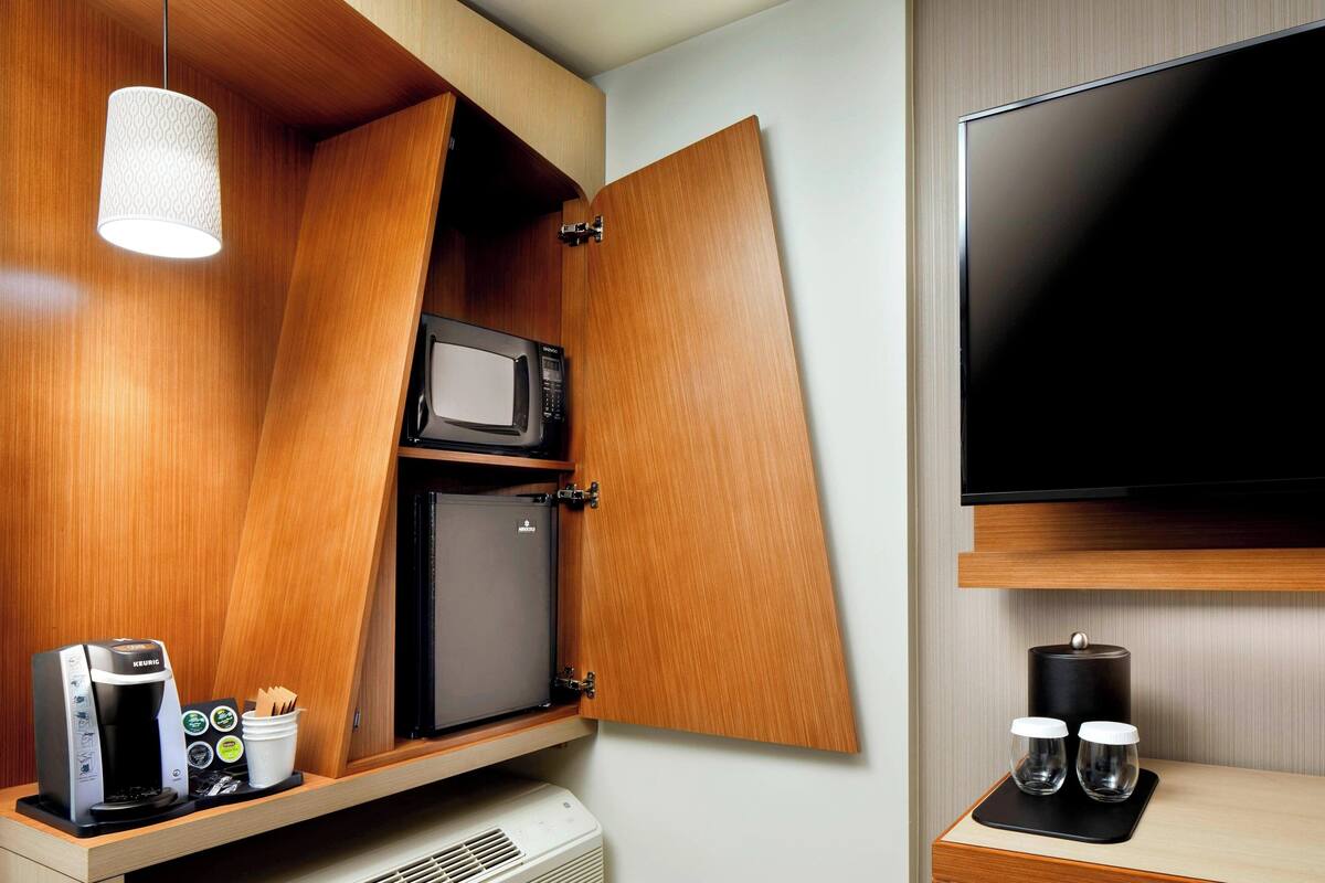 A compact kitchenette with a coffee machine, microwave, and a small refrigerator. A TV is mounted on the wall to the right.