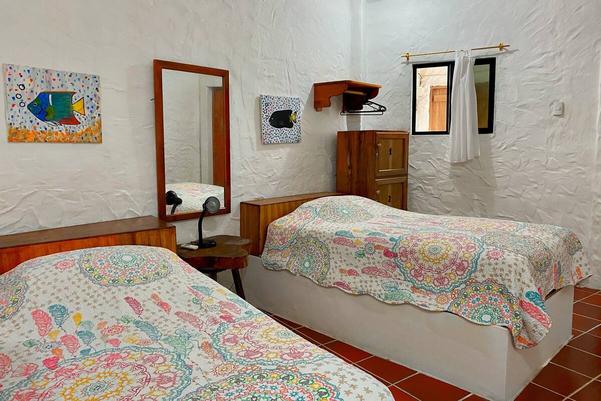 A bedroom with two beds, a wooden wardrobe, a mirror, a small table with a lamp, and a window with curtains.