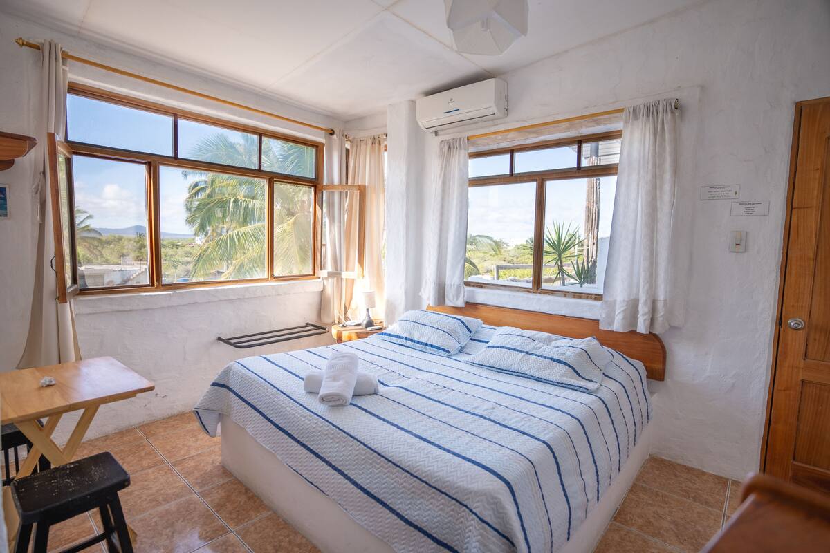 A bedroom with a bed, a desk, and a chair. There are windows with curtains and an air conditioning unit on the wall.