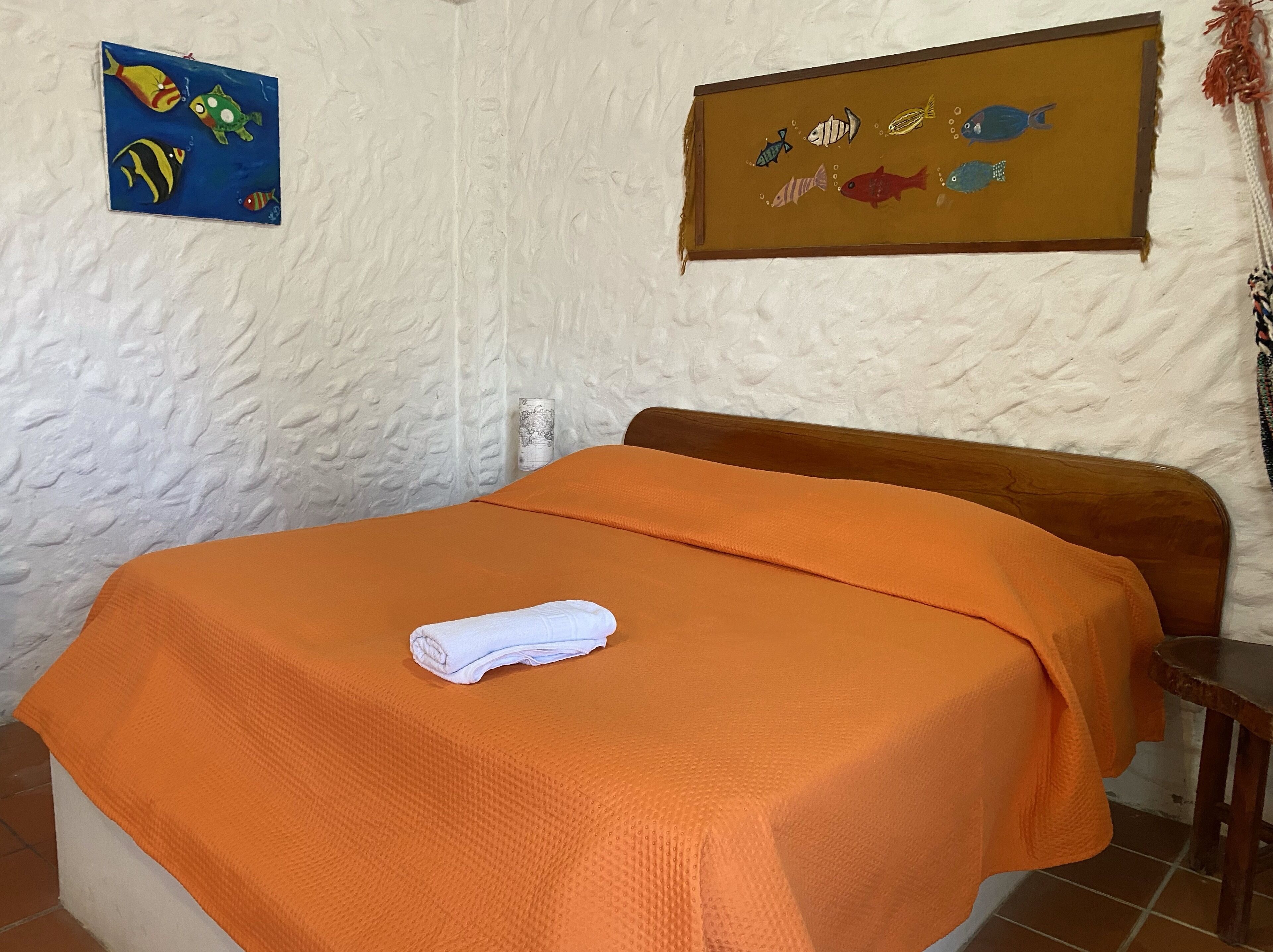 Ett sovrum med en sänggavel i trä, ett orange sängöverkast och en vit handduk på sängen. På väggarna hänger två dekorationer som föreställer fiskar.