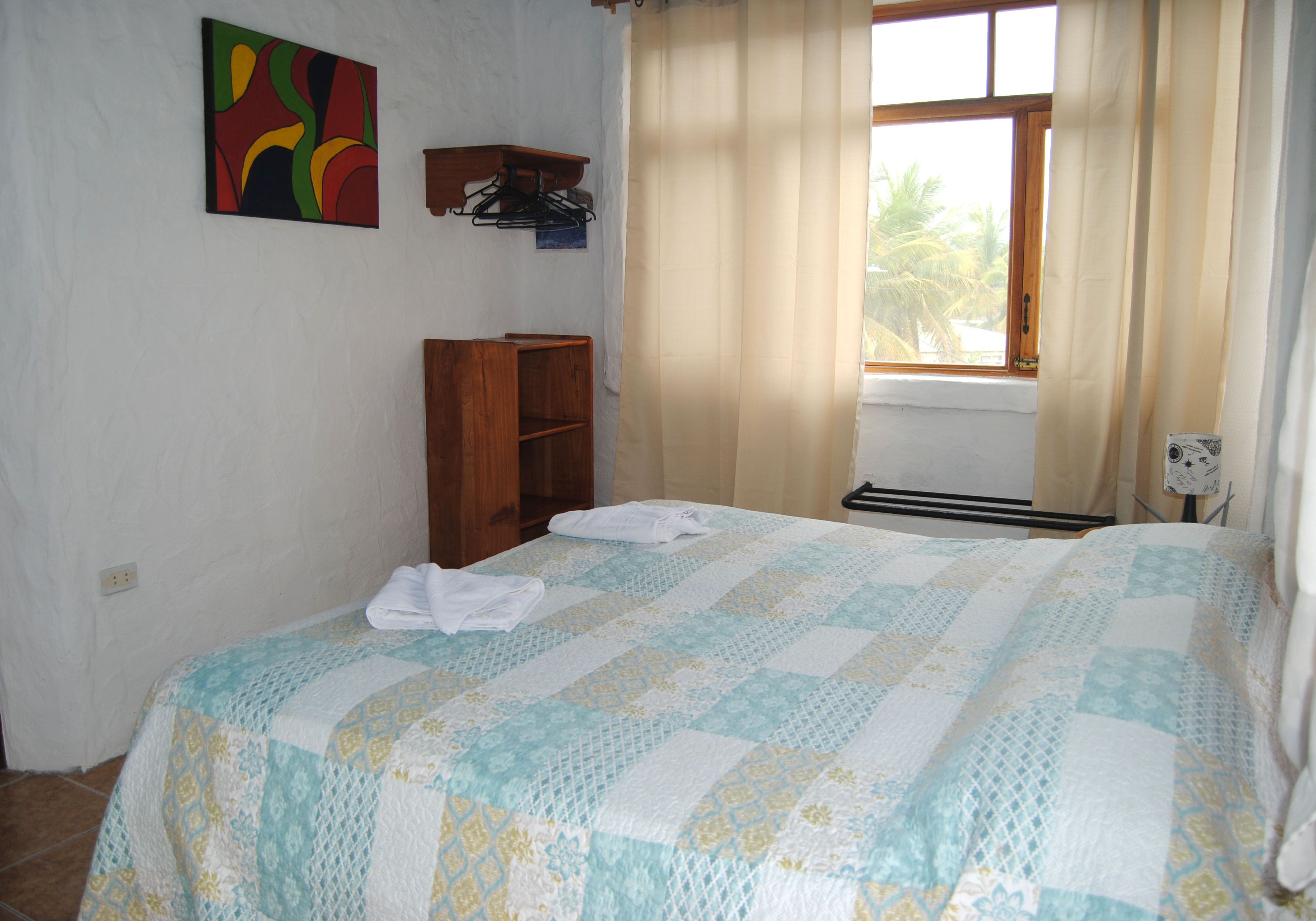 Een slaapkamer met een bed, een houten plank, een raam met gordijnen en een aan de muur hangend schilderij.