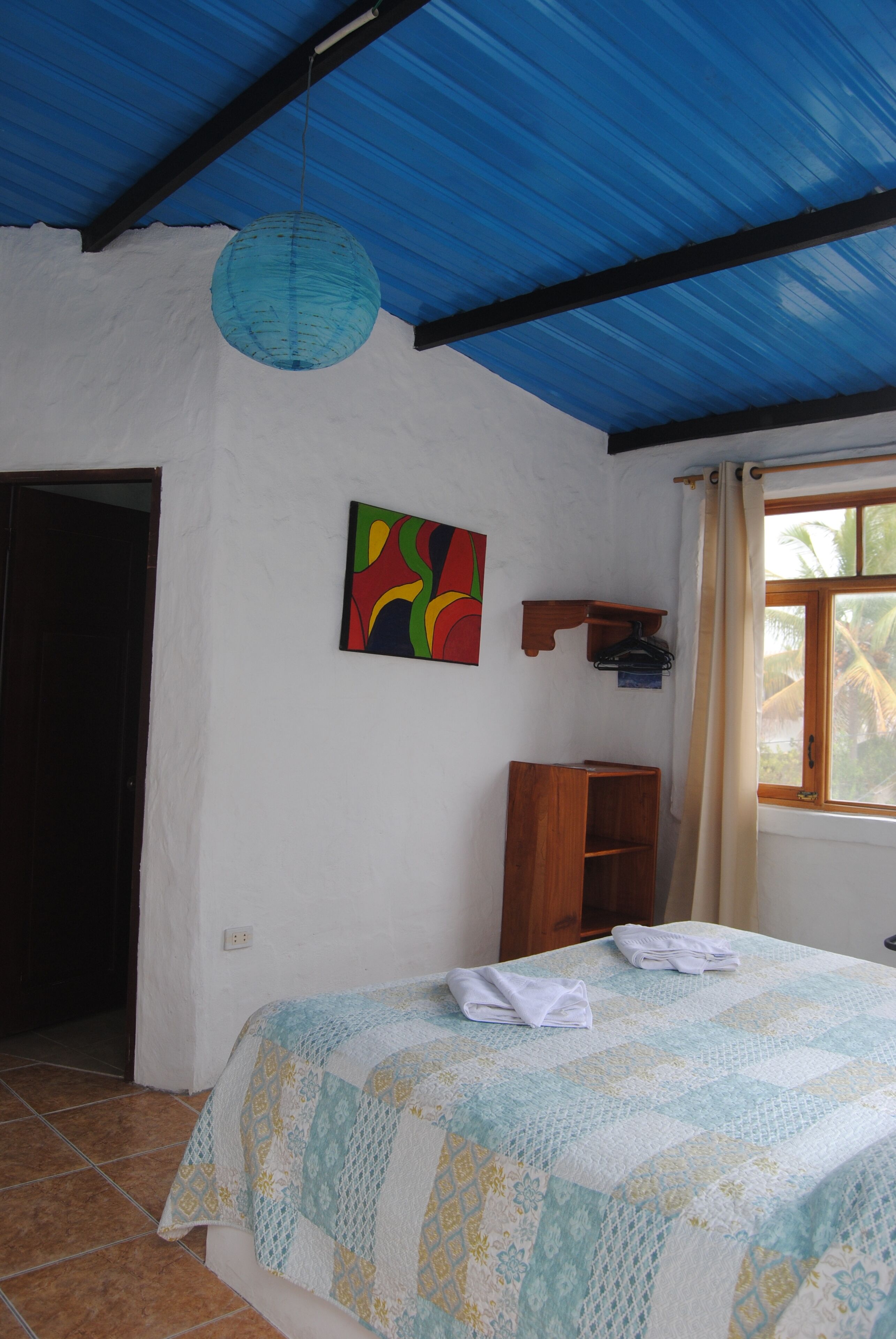 Un dormitorio con cama, una lámpara colgante, un cuadro de colores, un estante de madera y una ventana con cortinas.