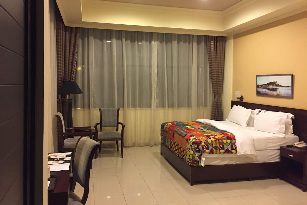 A hotel room with a bed, a desk with a chair, a lamp, a nightstand, and a painting on the wall.