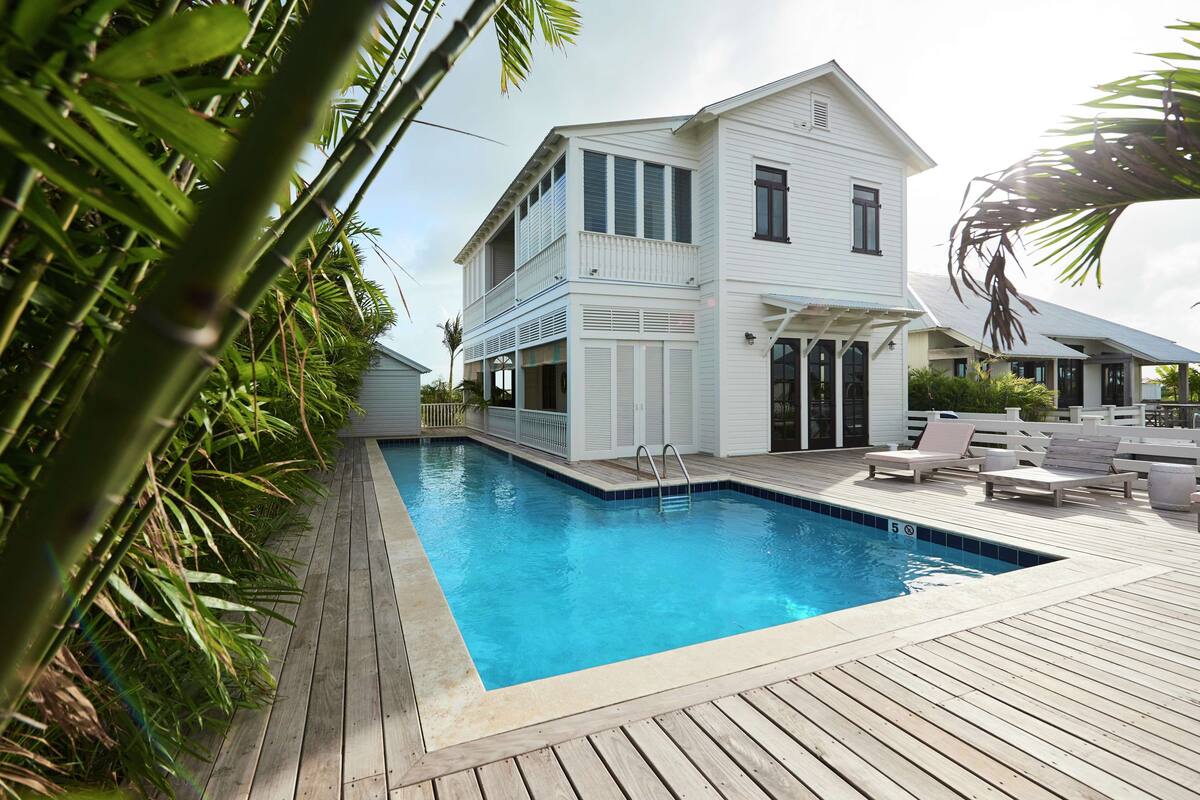 Une maison blanche à deux étages, dotée d’une piscine, d’une terrasse en bois et de mobilier d’extérieur.