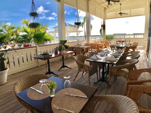 Un espacio de comedor al aire libre con mobiliario de mimbre, mesas dispuestas con servilletas y cubiertos blancos, y plantas colgantes.