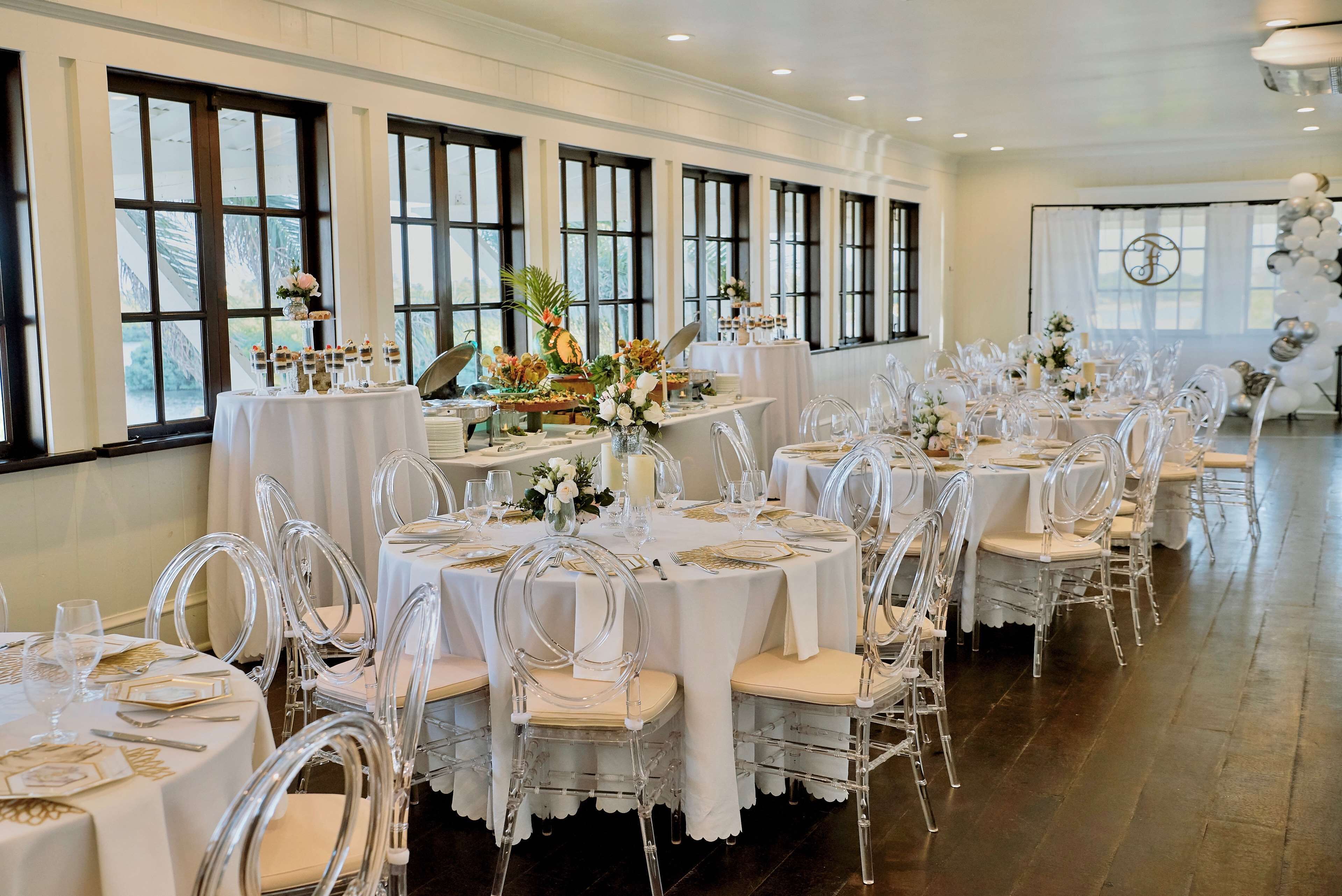 Uma sala de banquetes com mesas redondas, cadeiras transparentes e toalhas de mesa brancas.