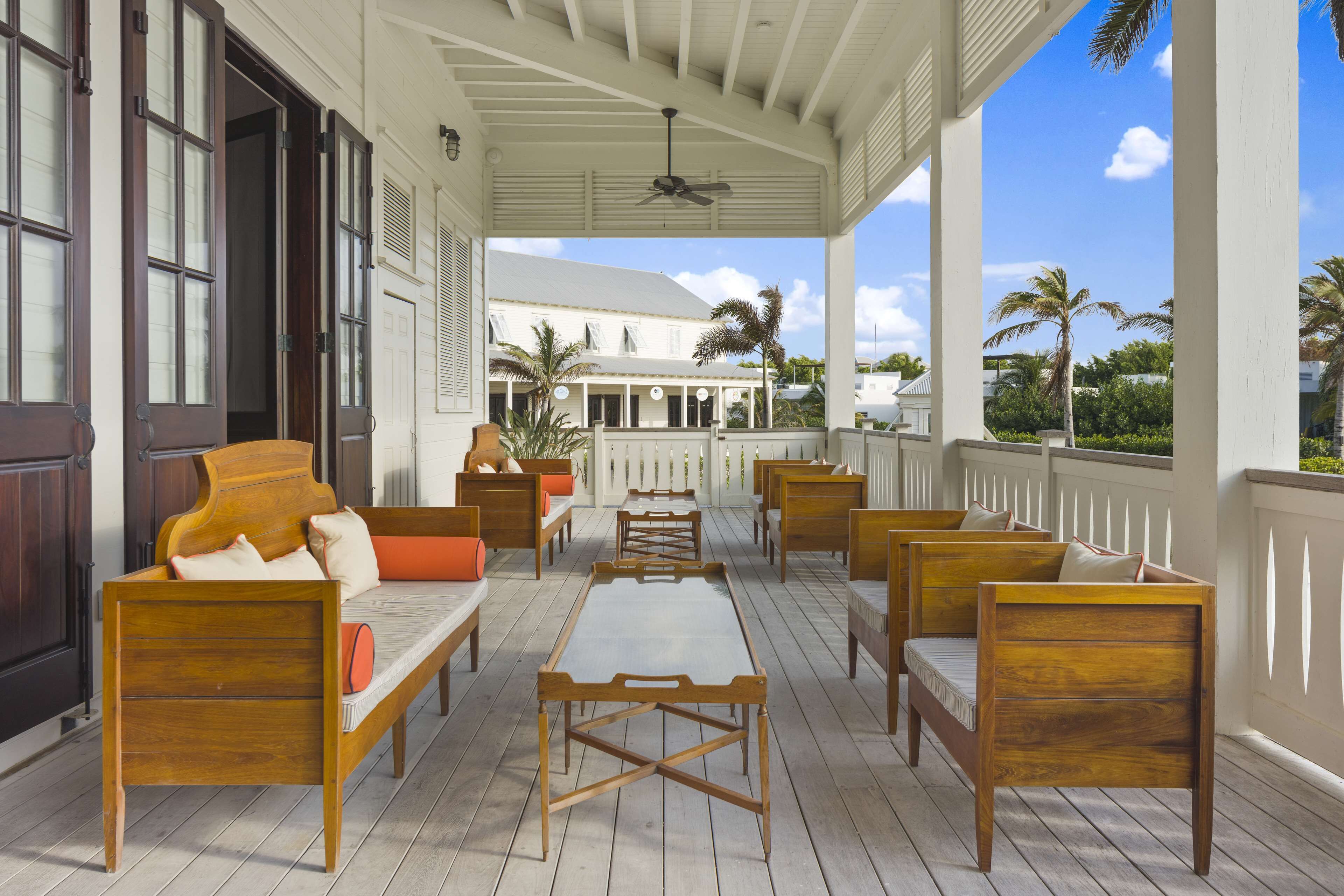 Eine überdachte Veranda mit Holzmöbeln, darunter Sofas und Stühle, und Blick auf ein weißes Gebäude im Hintergrund.
