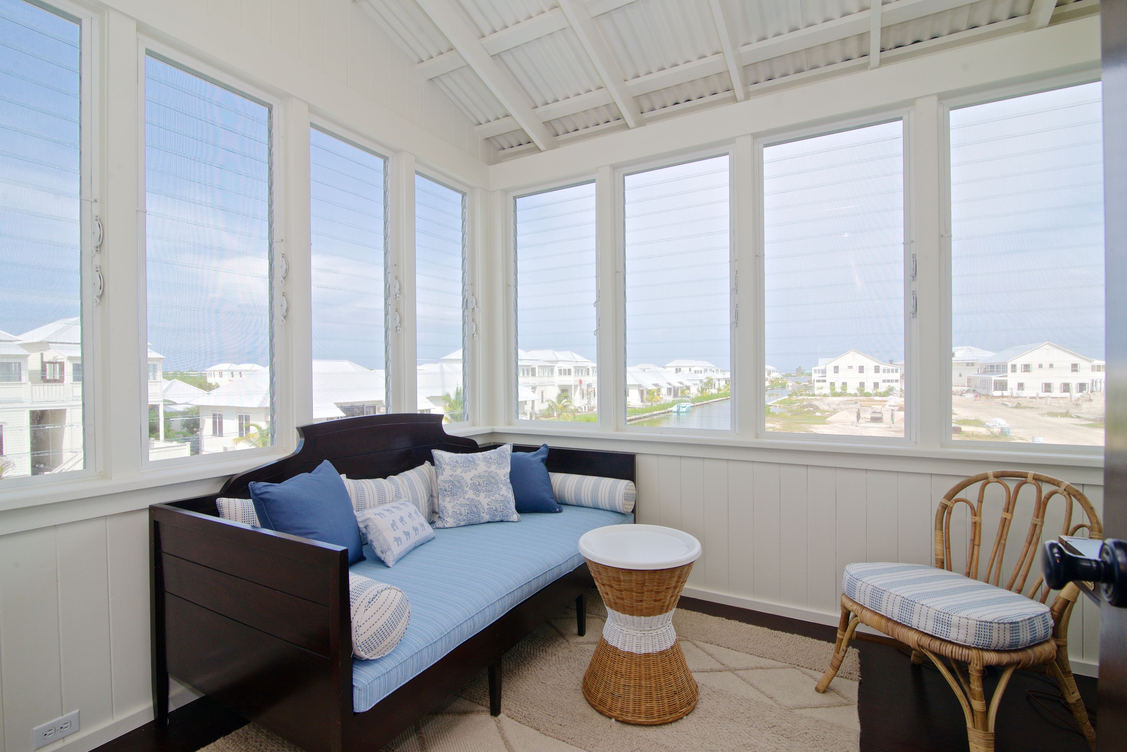 Una habitación con una cama, una silla de mimbre y ventanas amplias con vistas a edificios.