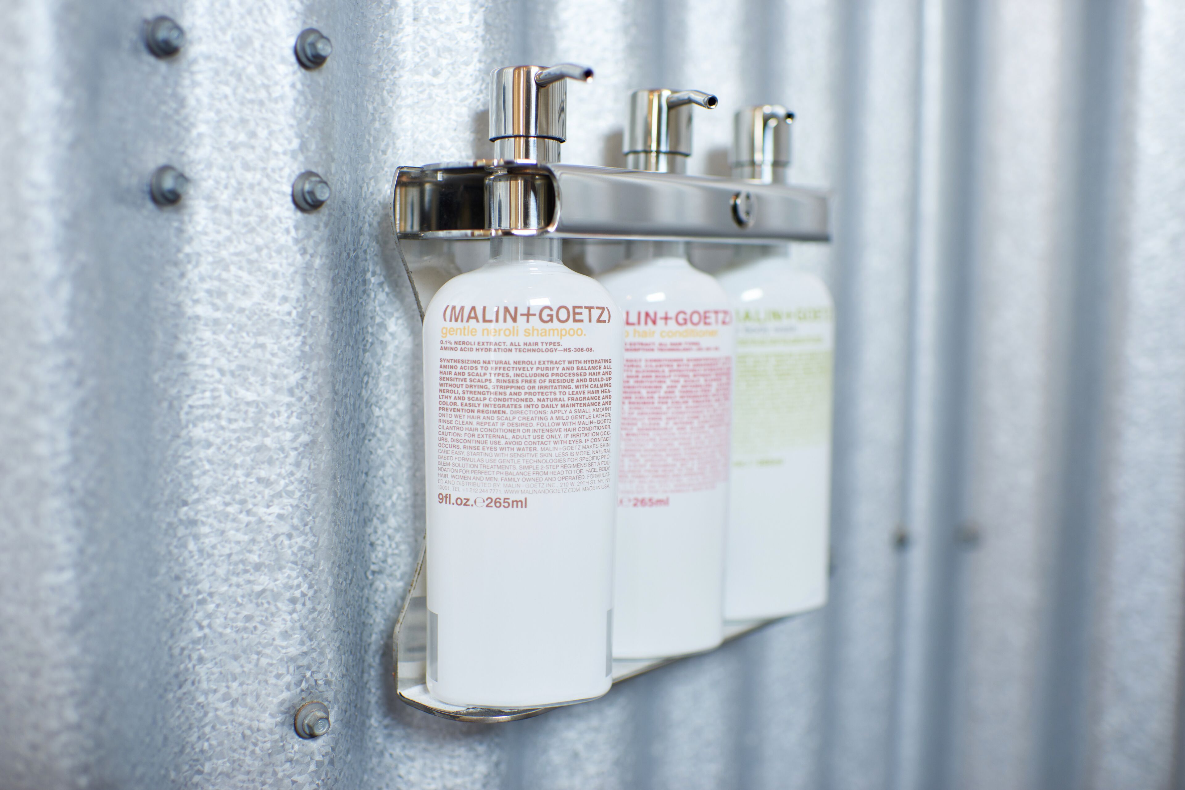 Una parete metallica con tre flaconi bianchi di shampoo Malin+Goetz.