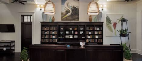 Un área de recepción con un mostrador de madera oscura, dos lámparas grandes adosadas a la pared, una estantería llena de libros y un cuadro de un pájaro.