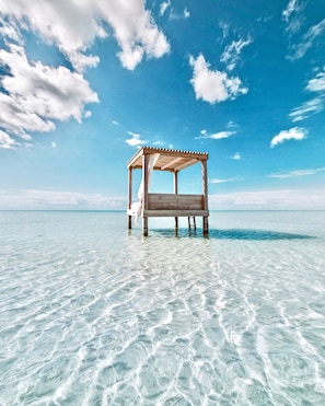 A wooden beach hut with a canopy, situated on a clear blue sea with shallow water.