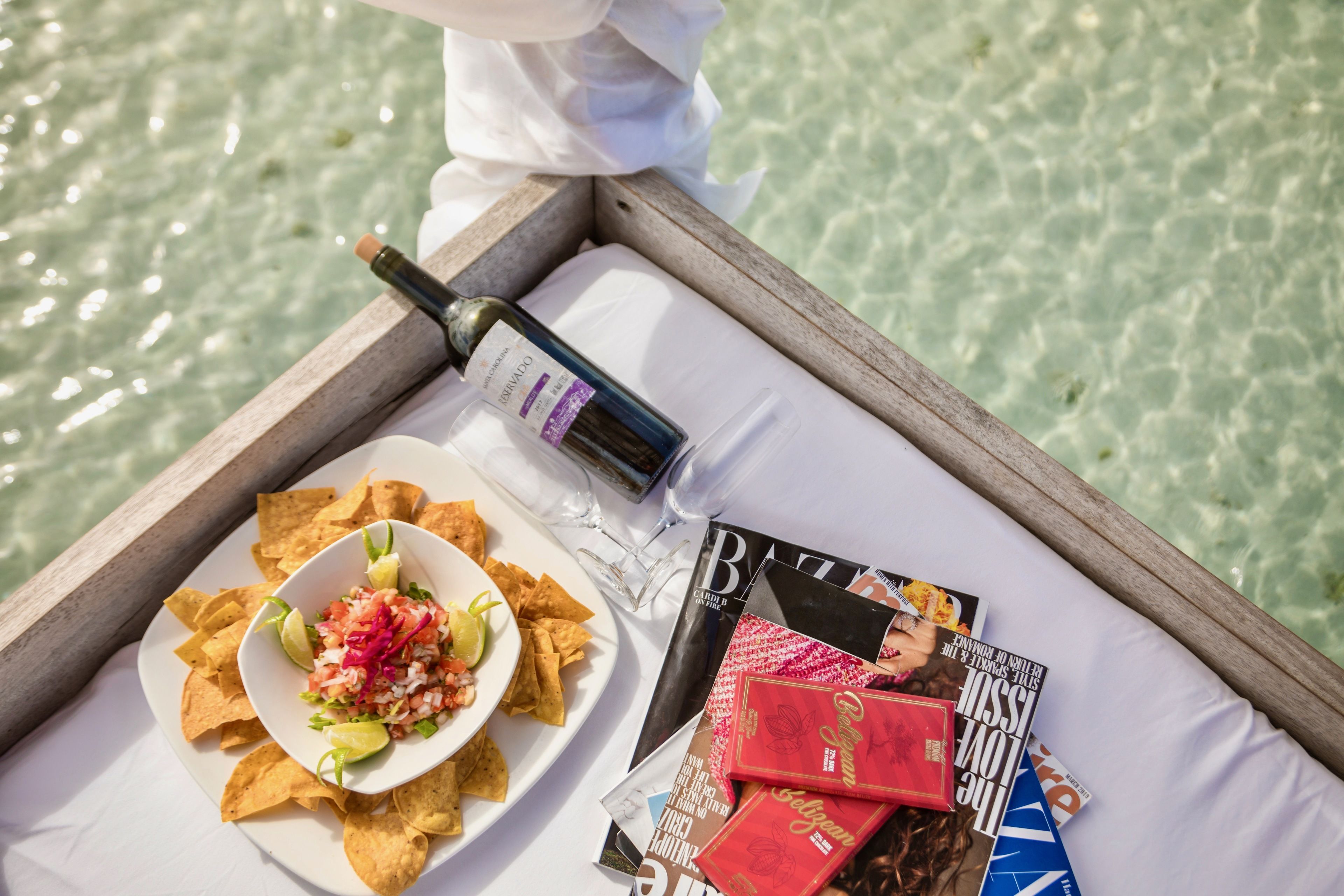 A tray with a plate of chips and salsa, a bottle of wine, and magazines on a white surface by a body of water.