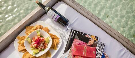 A tray with a plate of chips and salsa, a bottle of wine, and magazines on a white surface by a body of water.