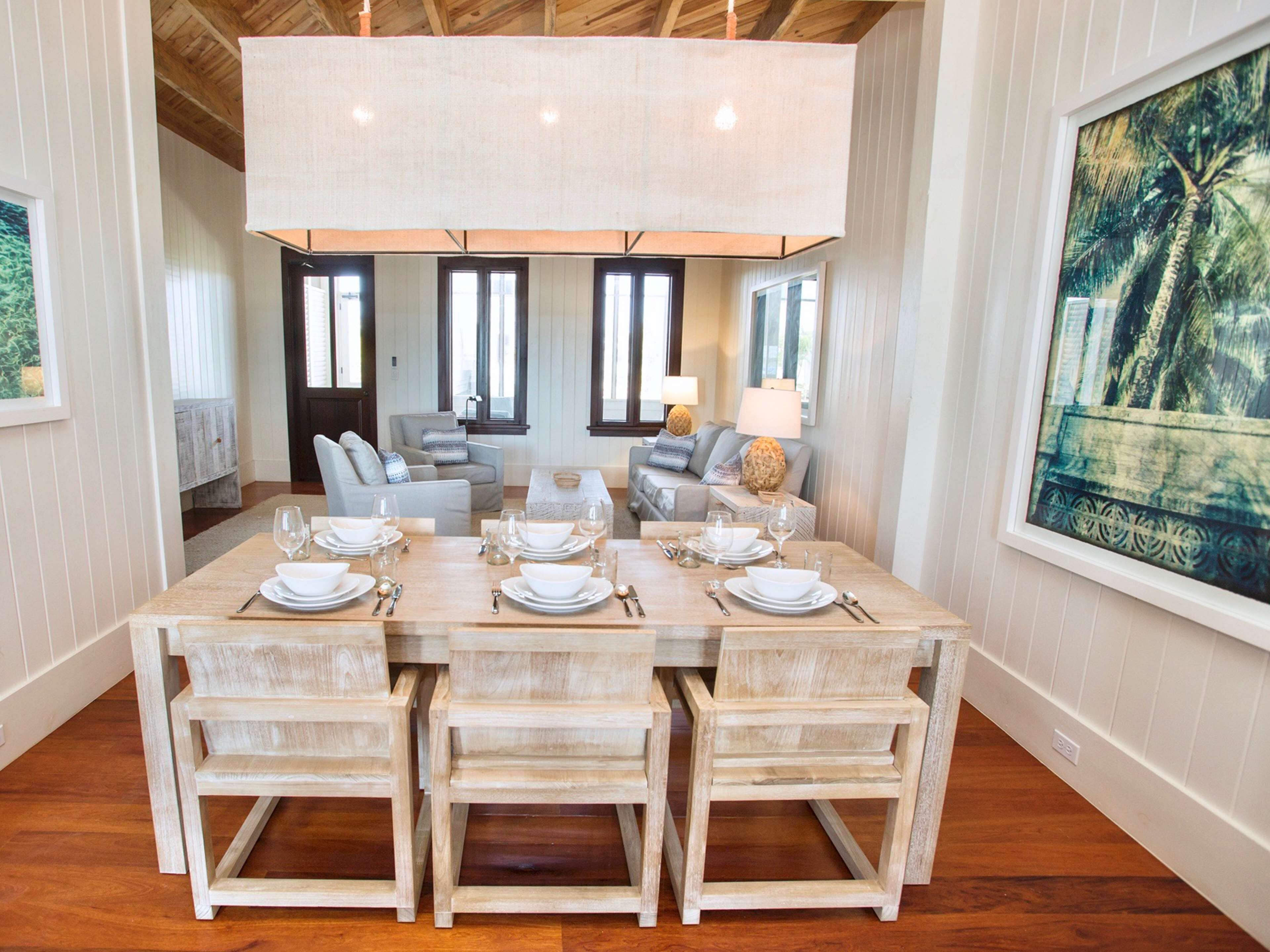 Un comedor con una mesa de madera preparada para seis personas, rodeada por cuatro sillas. Se observan dos sofás y una mesa de centro al fondo.