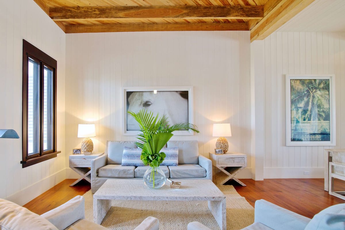 Un salon avec un plafond en bois, des murs blancs, un canapé gris, deux tables d’appoint avec des lampes, une table basse et un tableau encadré au mur.