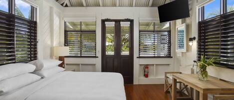 A bedroom with a bed, a desk, a chair, a television, and a window with shutters.