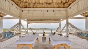 Eine Holzterrasse mit zwei Massagetischen, beschriftet mit 'Sierra Comfort', unter einem Reetdach mit Blick auf einen Strand.