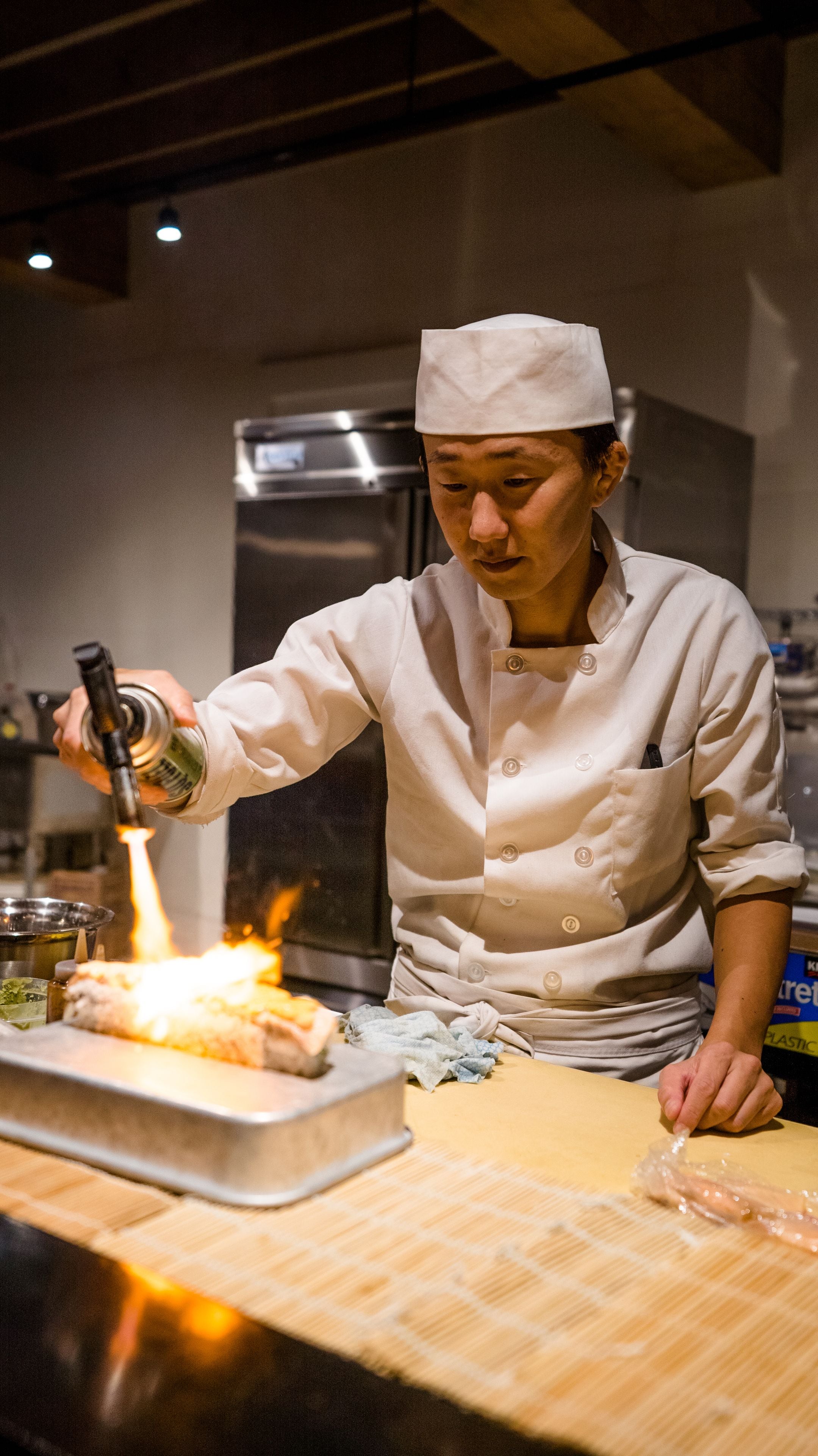 A chef in a white uniform with a hat, pouring a liquid from a bottle onto a pan on a stove.