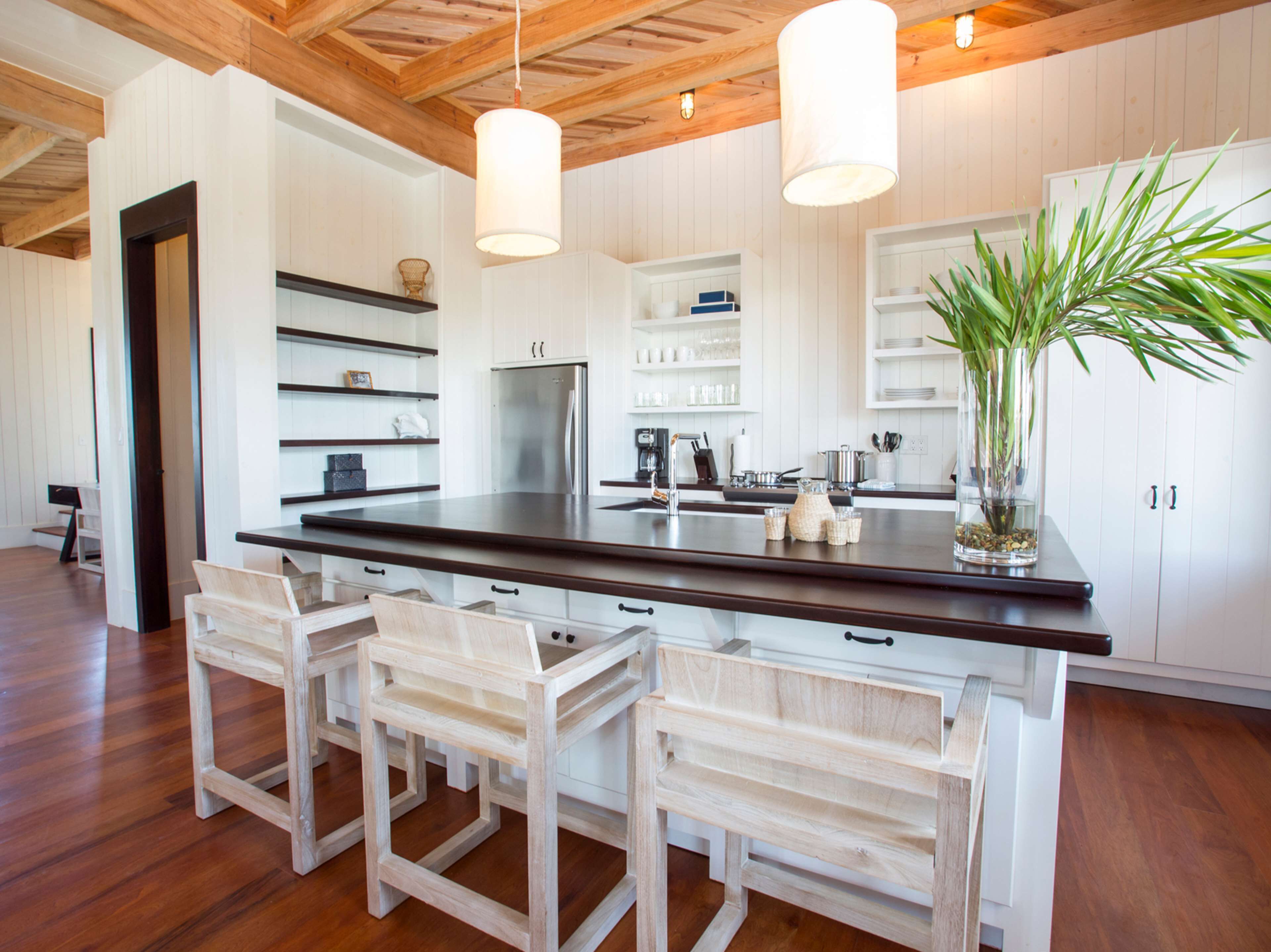 A modern kitchen with a central island, wooden chairs, white cabinetry, and pendant lighting.