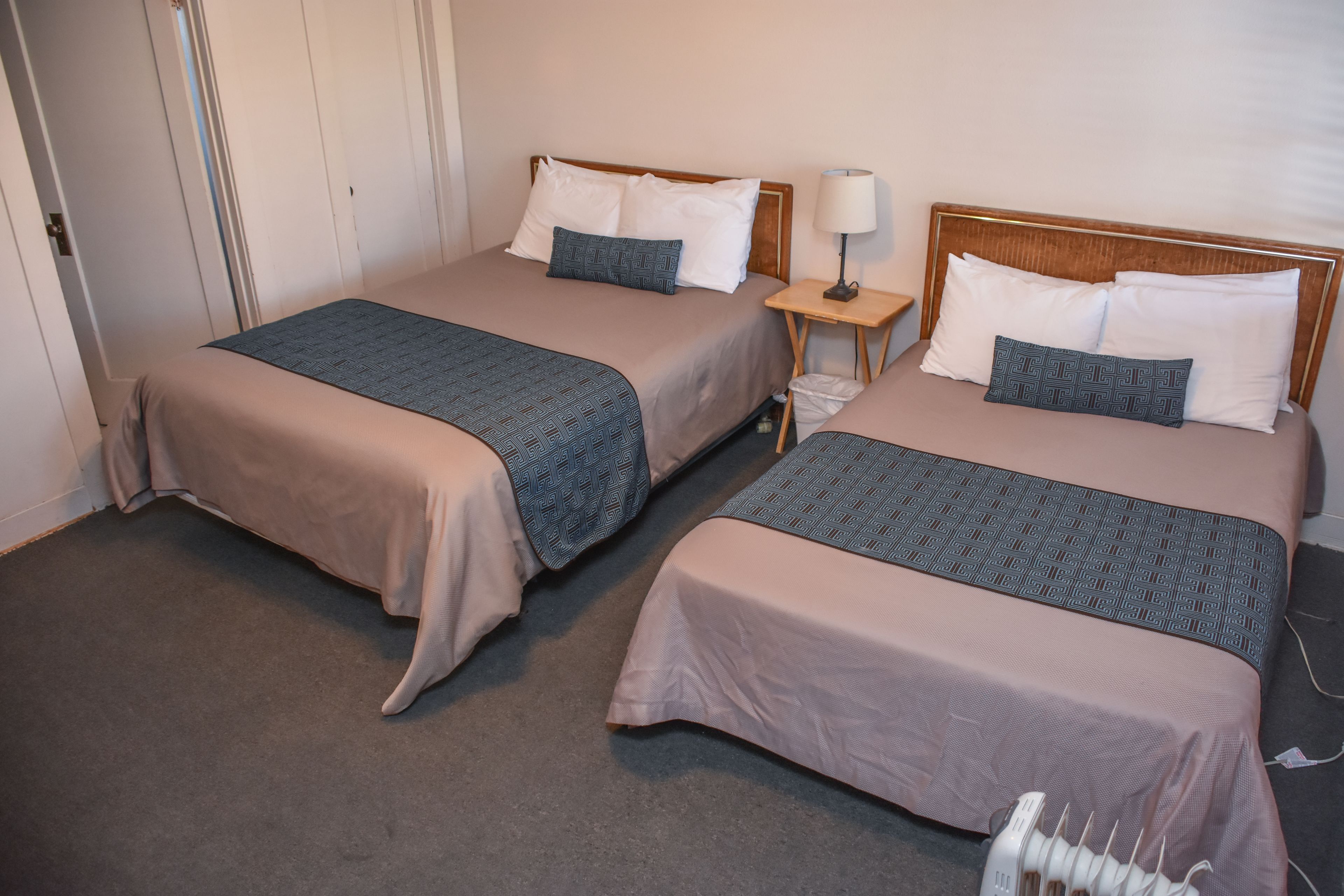 Two twin beds with beige bedding and patterned throw blankets, a bedside table with a lamp, and a white radiator.