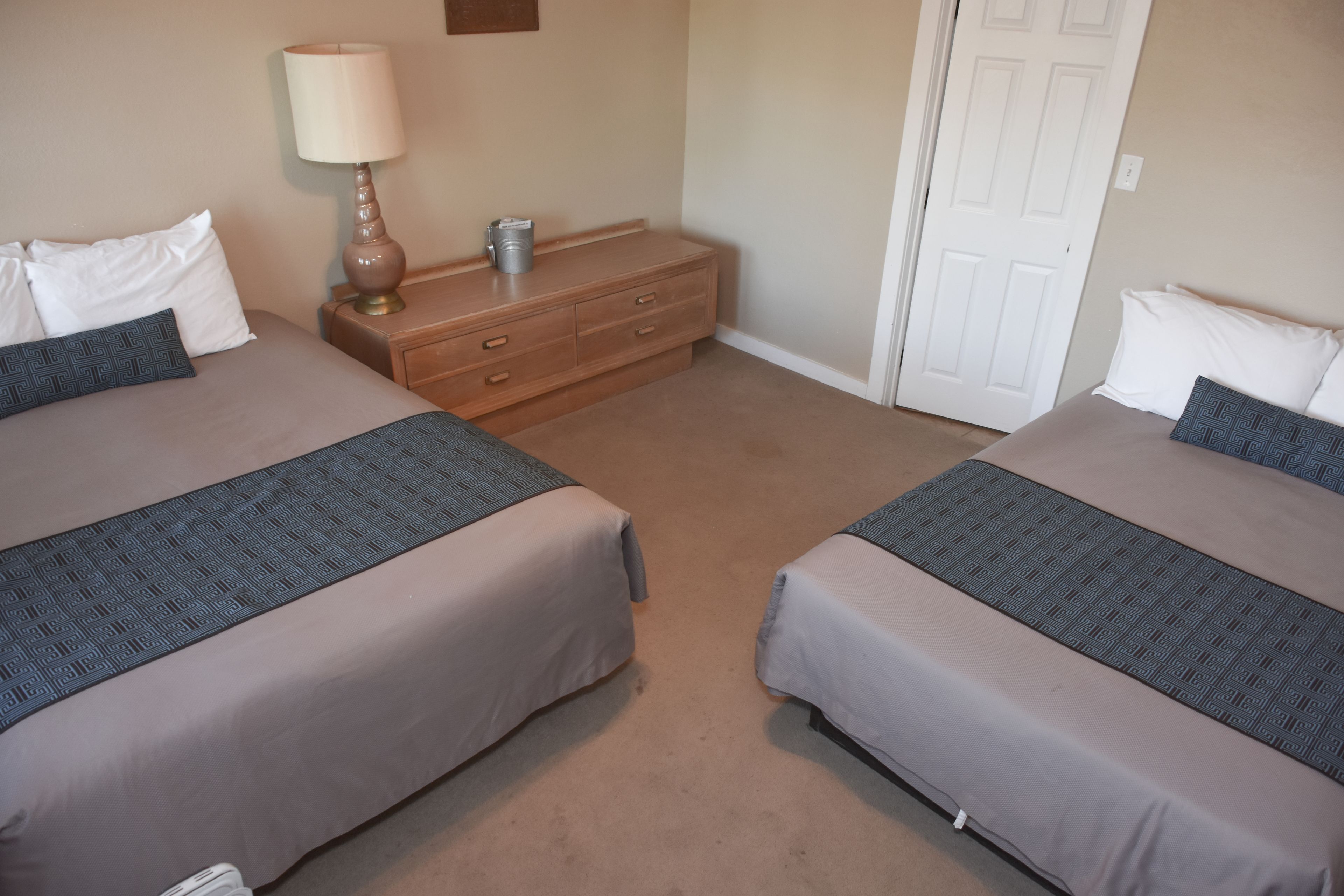 A hotel room with two beds, a nightstand with a lamp, a dresser, and a door.