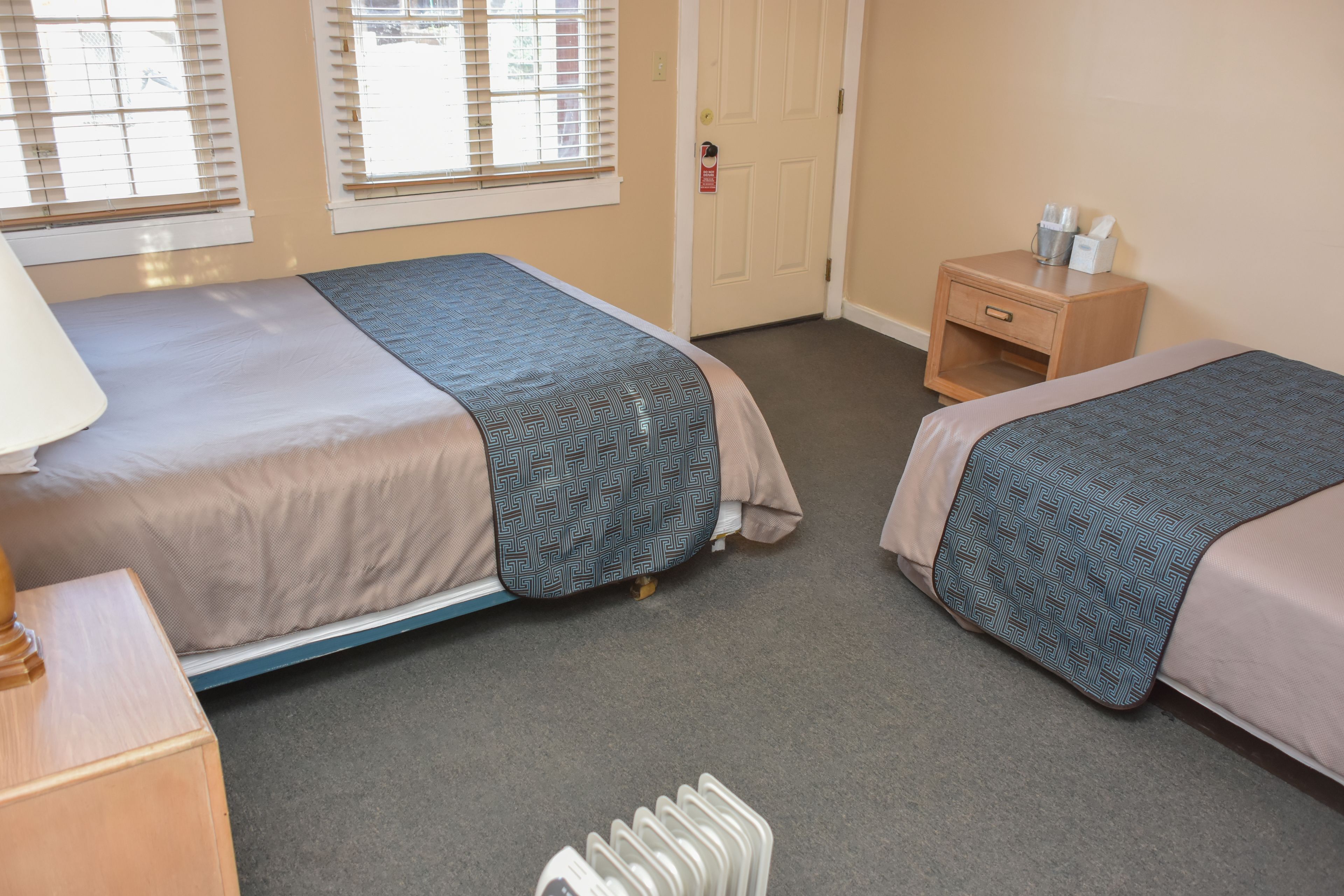 A hotel room with two beds, a nightstand, a lamp, and a door.