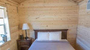 A wooden cabin room with a bed, a nightstand, a lamp, a ceiling fan, and a window. - Panamint Springs Resort (Panamint Springs)