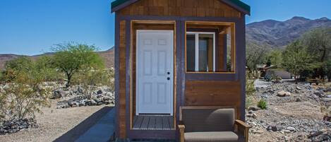 Una pequeña cabaña de madera con puerta y ventana, un banco en el exterior y un paisaje desértico con montañas al fondo.