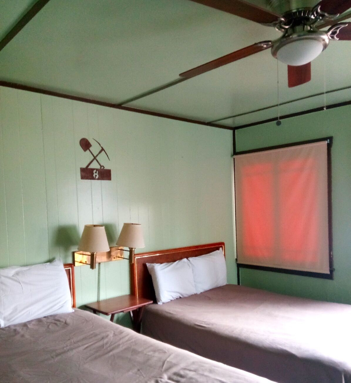 Una habitación con dos camas, un ventilador de techo, una mesita de noche con lámparas y una ventana con cortina roja.