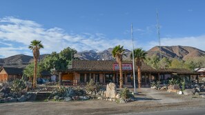 A rustic building with a sign advertising pizza, burgers, and salads. It features a stone facade, palm trees, and a mountainous backdrop.