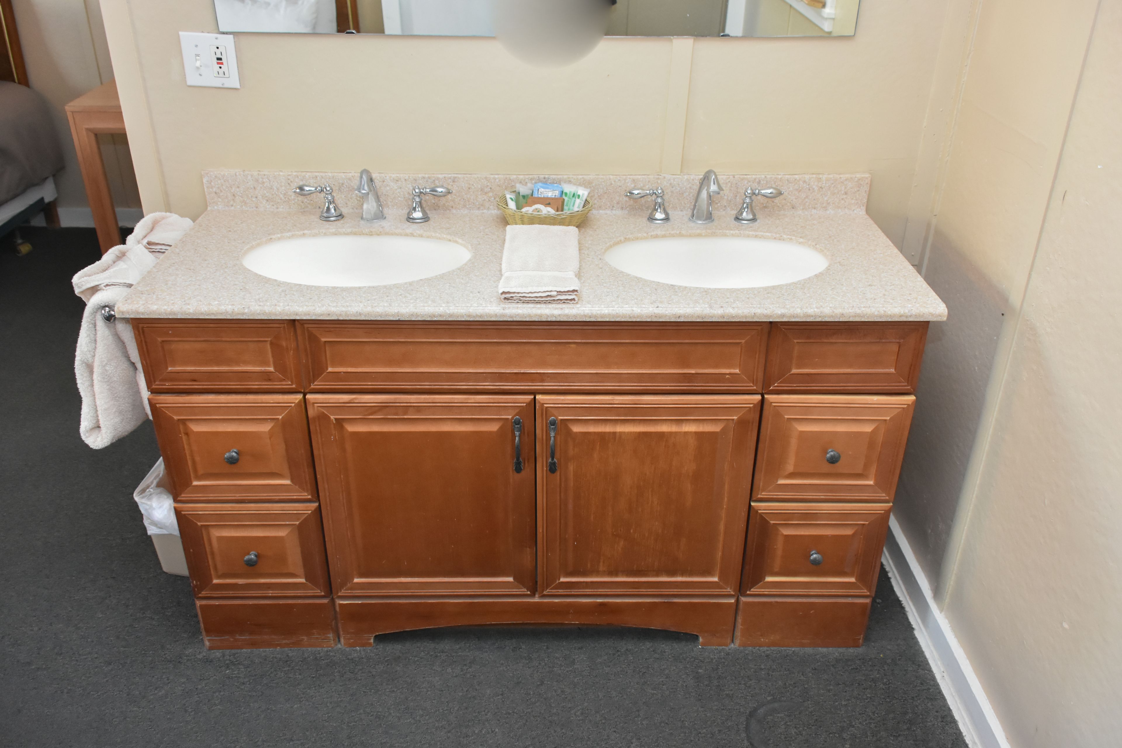 A wooden vanity with two sinks, a mirror, and a basket of toiletries.
