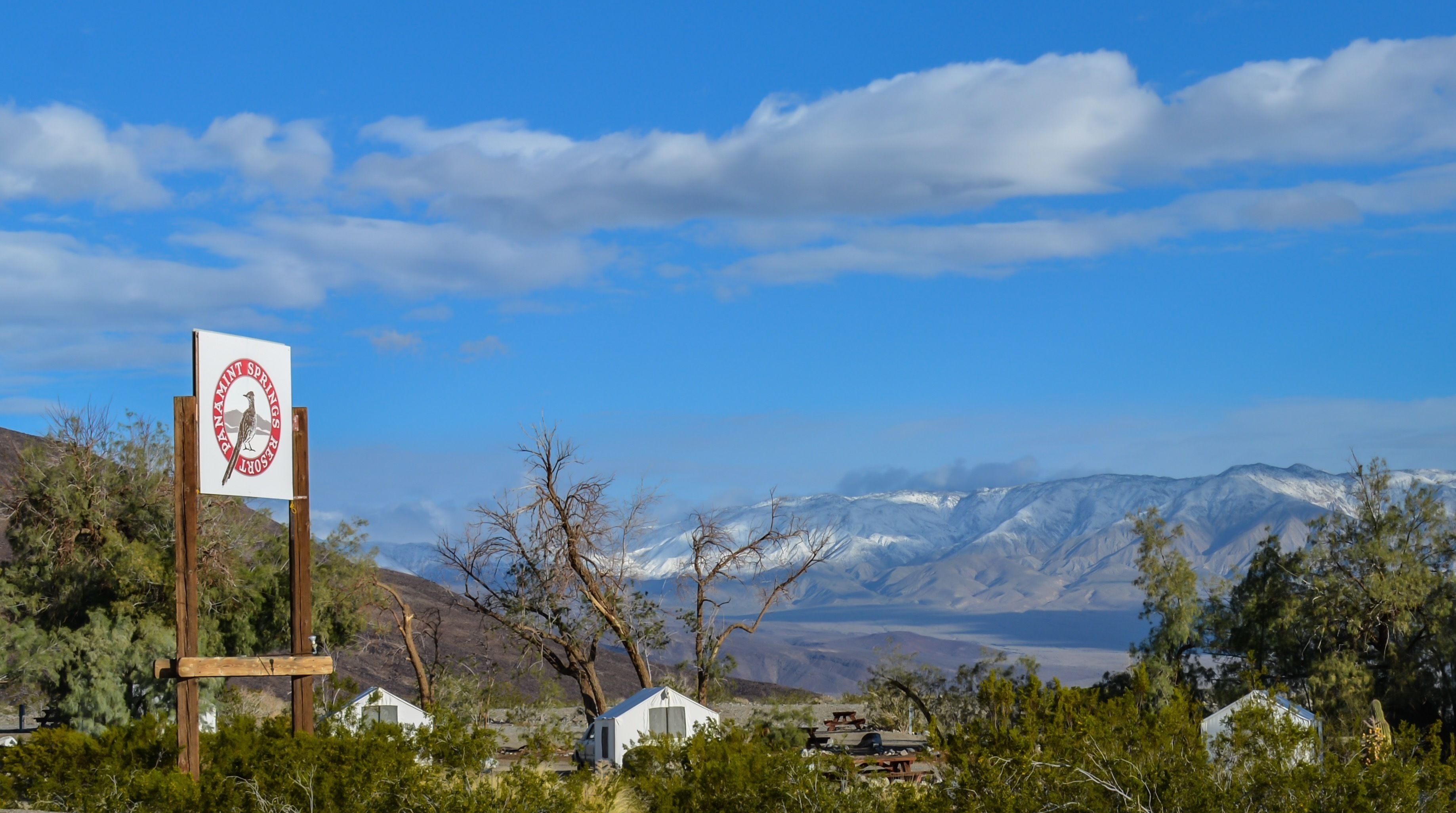A sign with a bird and text, surrounded by trees and a mountainous landscape.