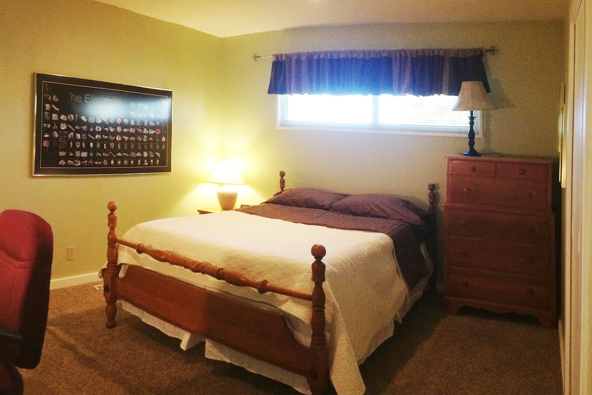 A bedroom with a bed, desk, chair, dresser, and a wall-mounted lamp.