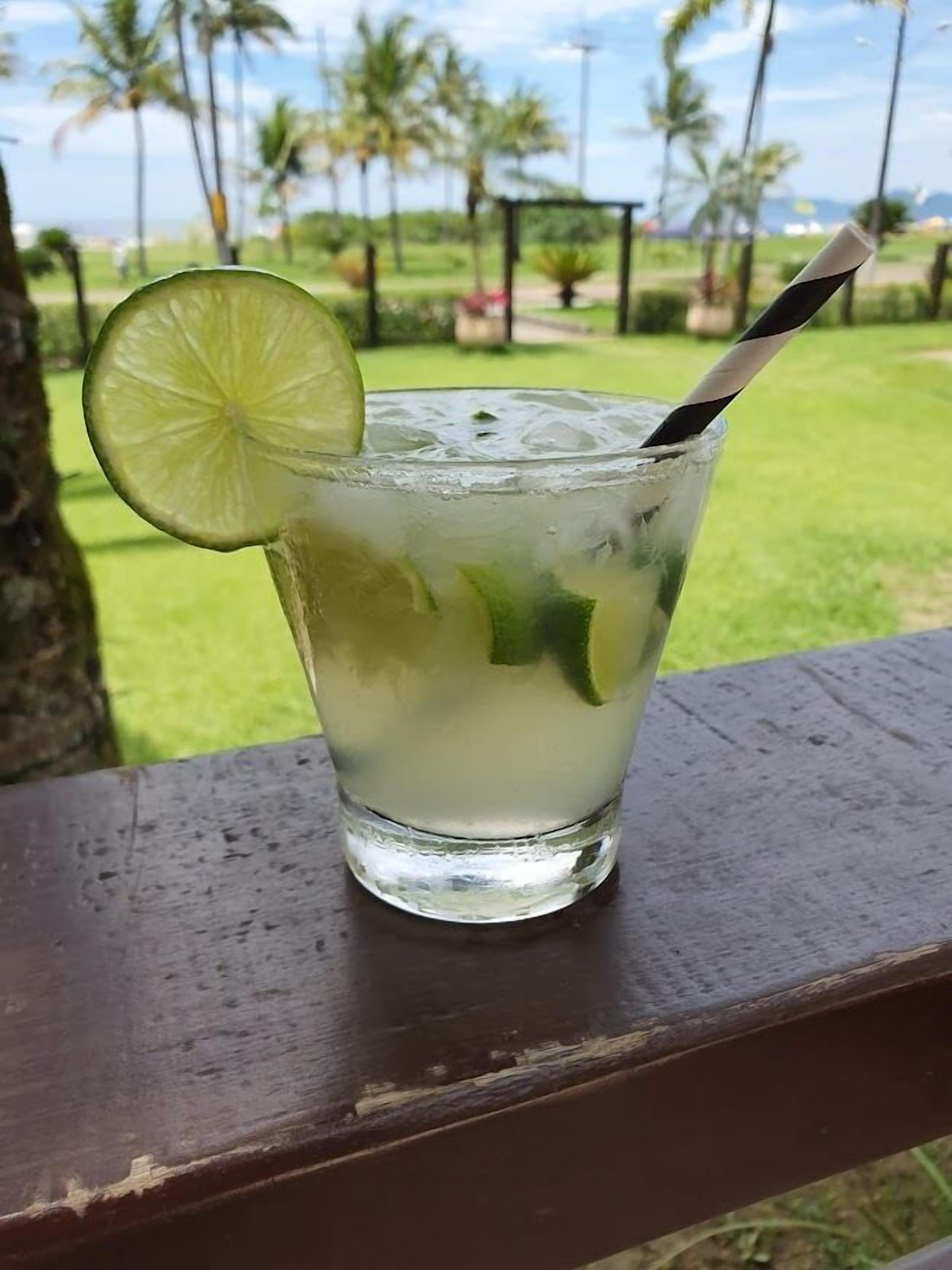 Un vaso de bebida helada con lima y menta, sobre una superficie de madera con vistas a palmeras y una zona verde al fondo.