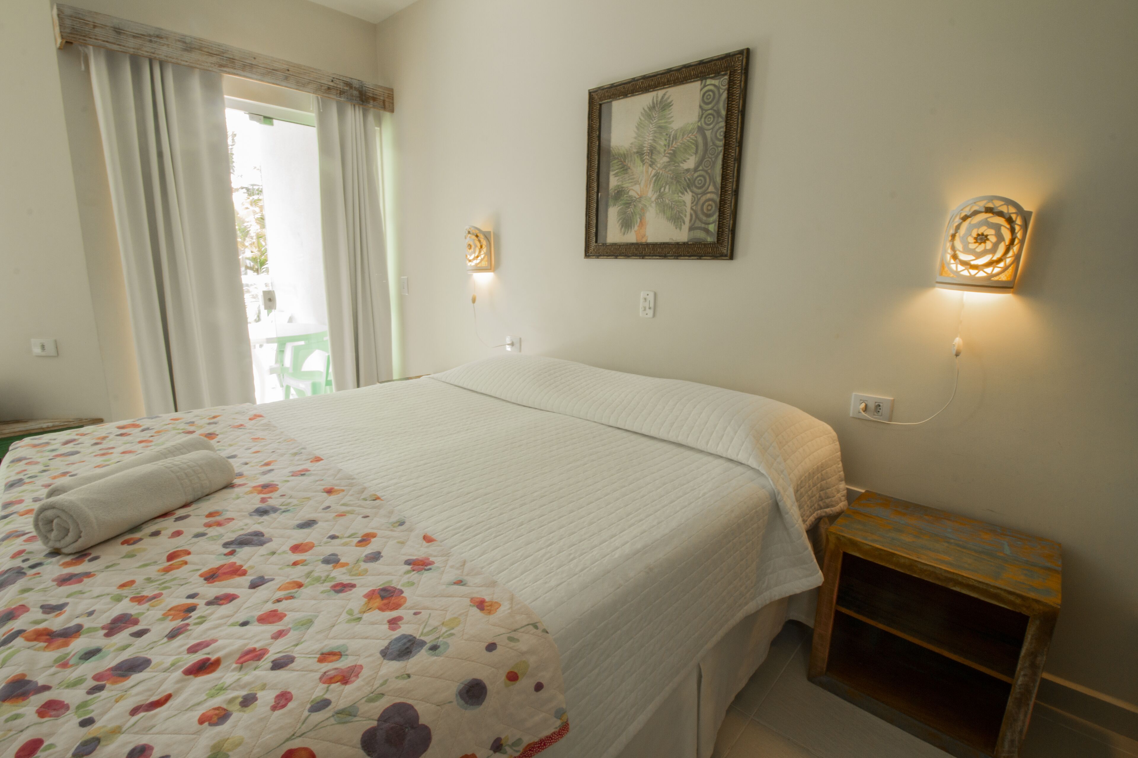 A neatly made bed with a floral-patterned bedspread, a small bedside table with a lamp, and a framed picture on the wall.