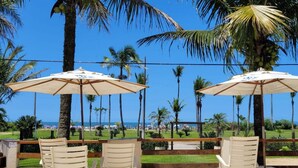 Een terras met witte stoelen en parasols, met uitzicht op een strand met palmbomen en een heldere lucht.