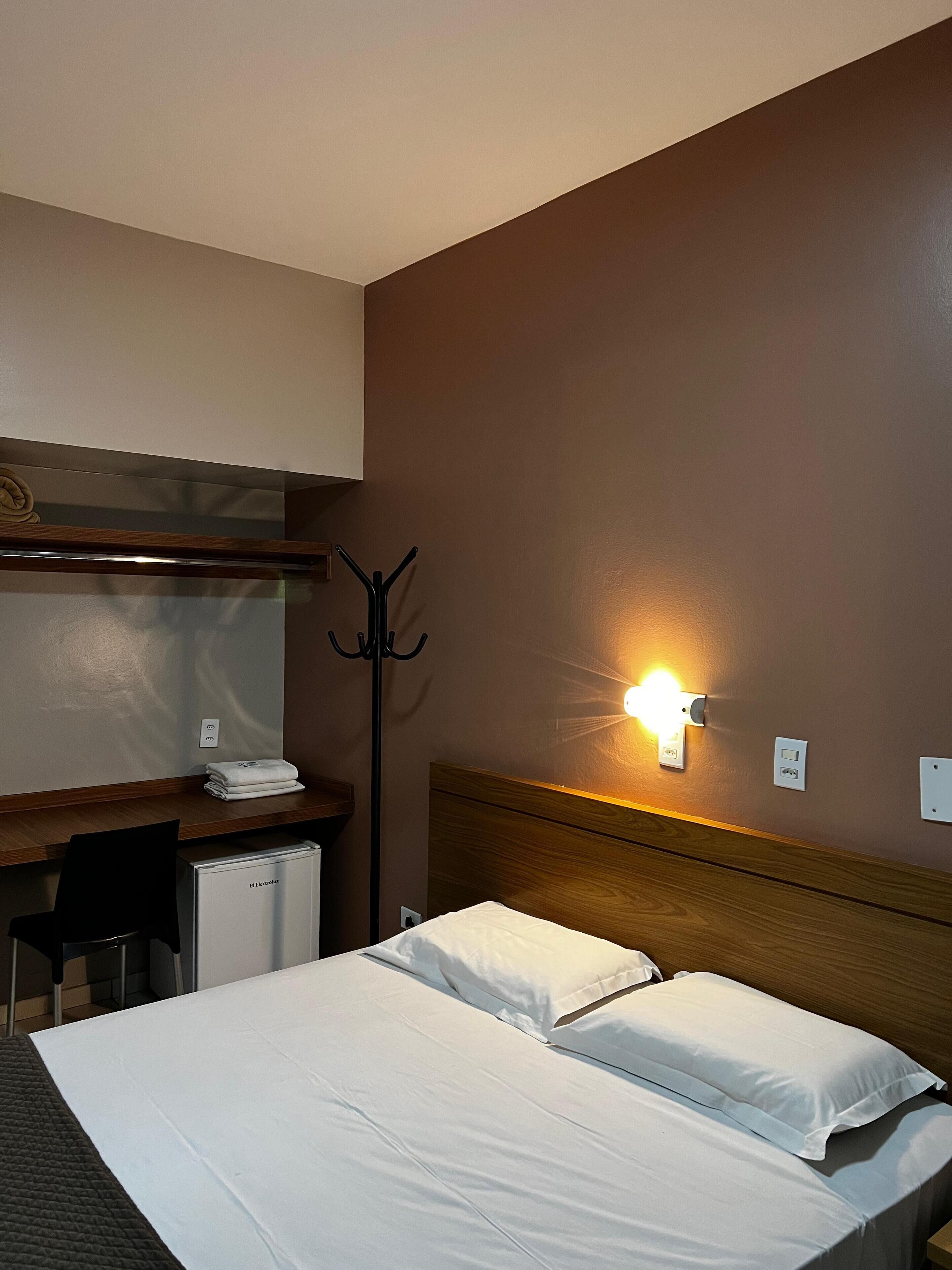 Quarto de hotel com cama, escrivaninha com cadeira, porta-casacos e um pequeno frigorífico.