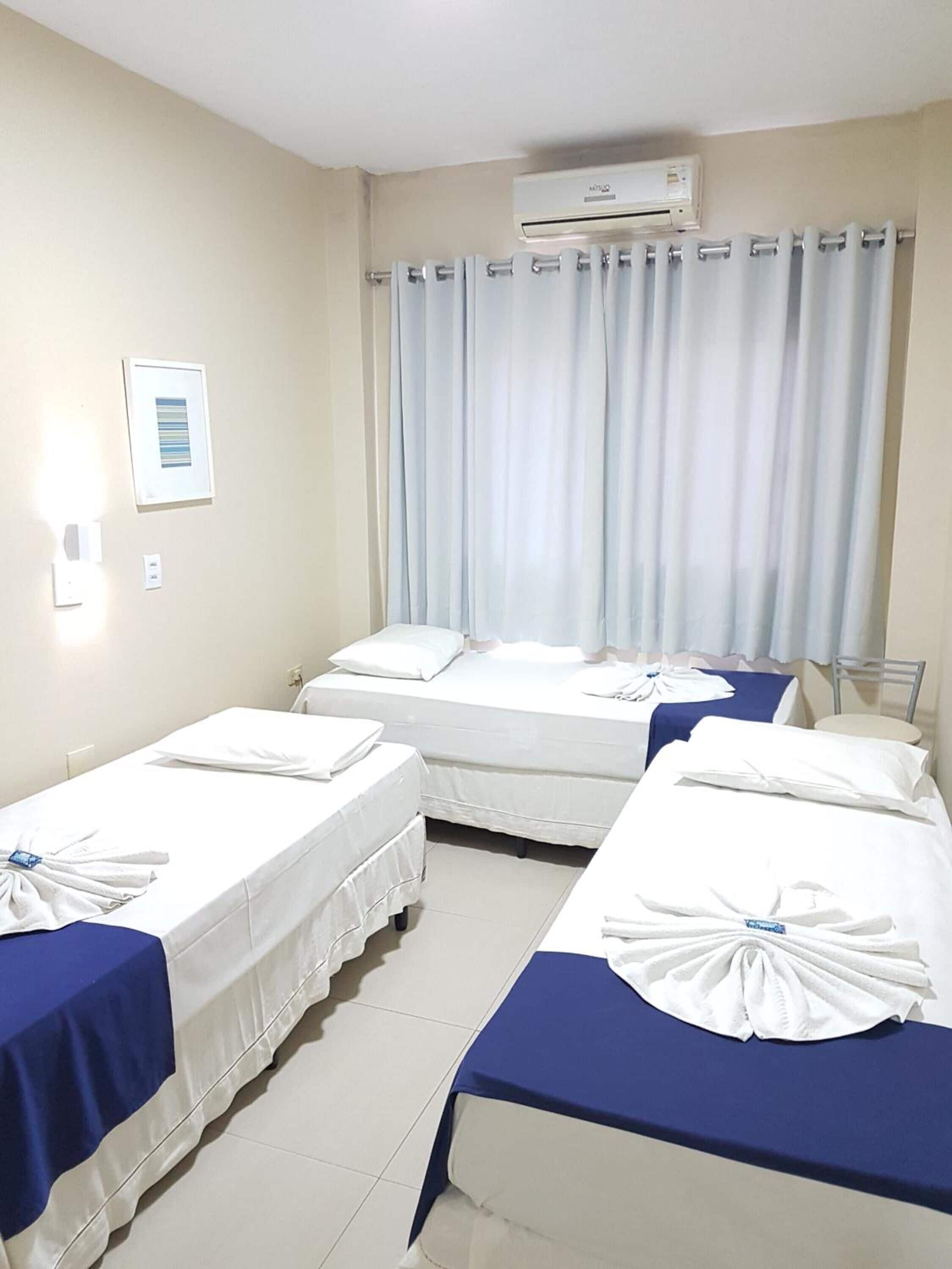 Hotellihuone, jossa on kaksi erillistä sänkyä, suuri ikkuna, jossa on verhot, ilmastointilaite ja seinällä oleva kehystetty taulu.