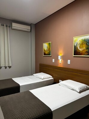 Habitación de hotel con dos camas individuales, aire acondicionado de pared, dos cuadros y una lámpara de noche.